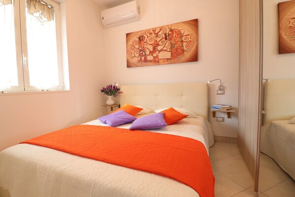 Quarto com cama grande, mesa de cabeceira, ar-condicionado na parede, um quadro e uma janela com cortinas.
