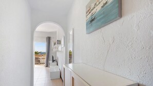 Un pasillo con pared blanca, un cuadro de una escena costera y vista a una habitación con bañera y mesa.