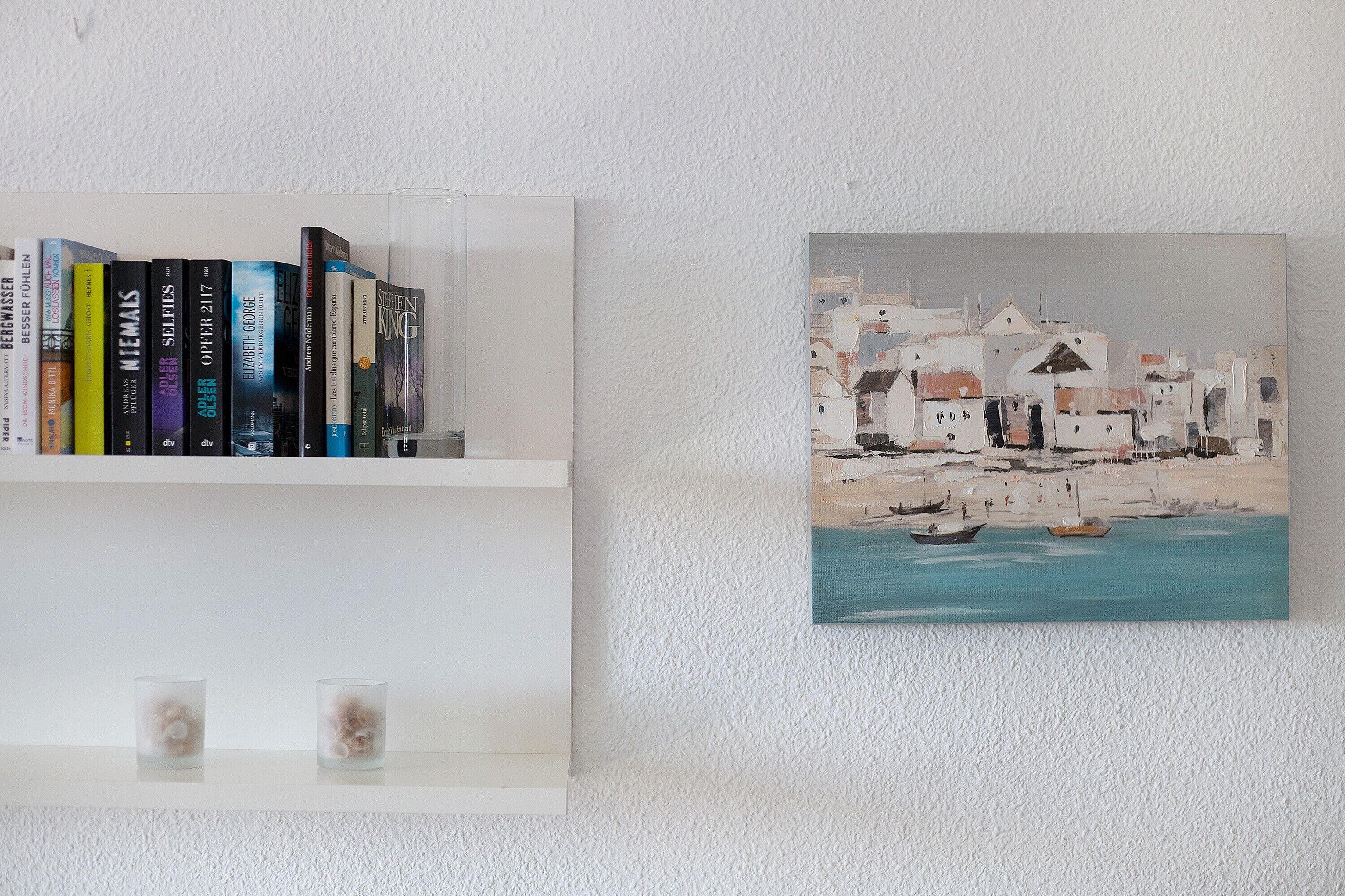 A bookshelf with books and a painting of a coastal scene.