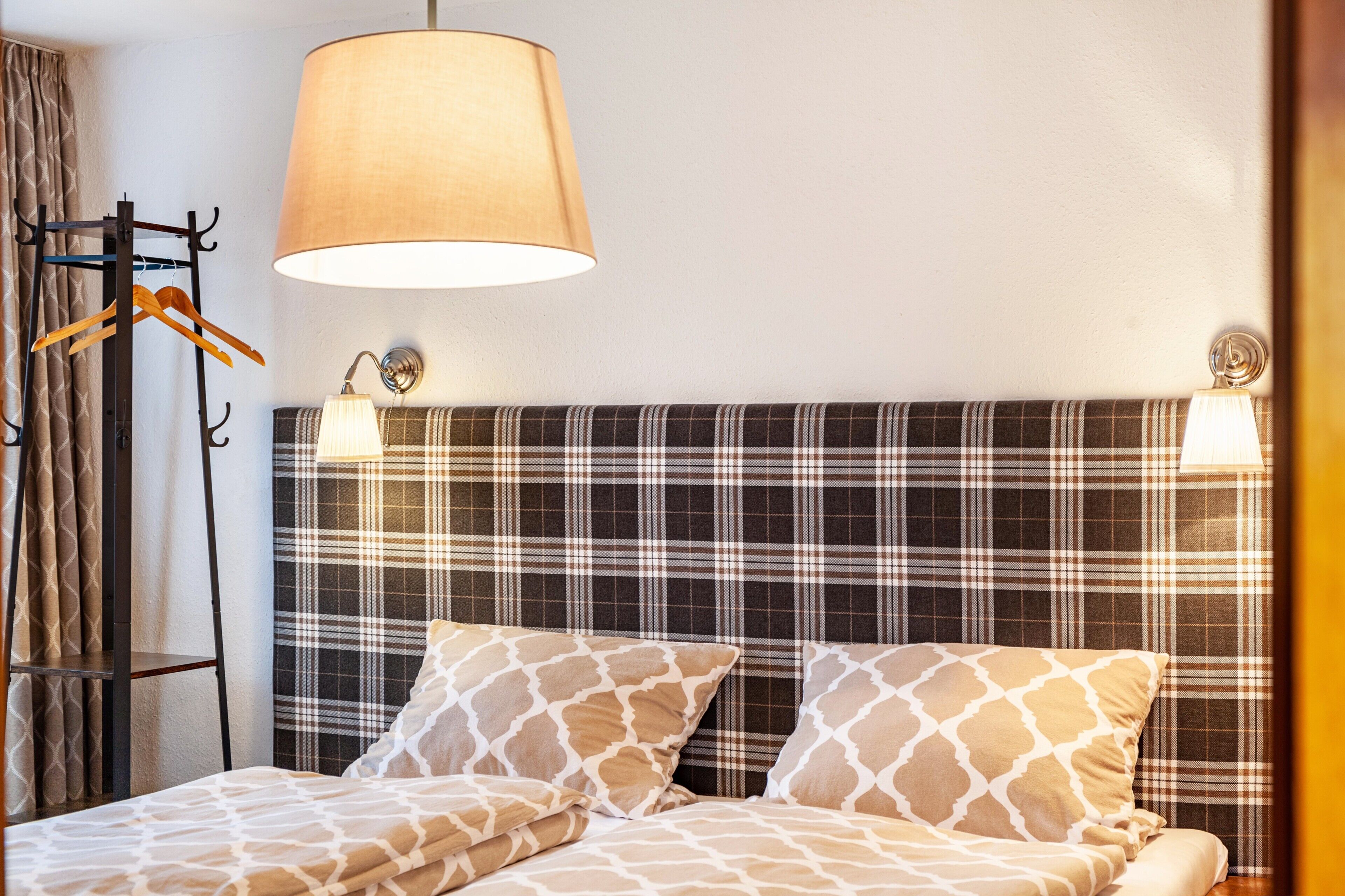 Une chambre d’hôtel avec un lit, une tête de lit à motifs écossais, une lampe suspendue et un porte-manteau avec un manteau.