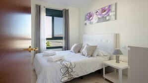 Une chambre à coucher avec un lit, une table de chevet, une lampe, une fenêtre avec des rideaux et un tableau au mur.
