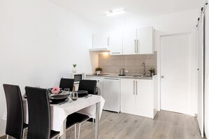 Een compacte keuken met witte kastjes, een eettafel met stoelen en een klein aanrecht met een spoelbak en een magnetron.
