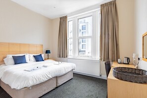 A hotel room with a large bed, a wooden headboard, a desk with a basket, and a window with curtains. - Brooklands Lodge (Bournemouth)