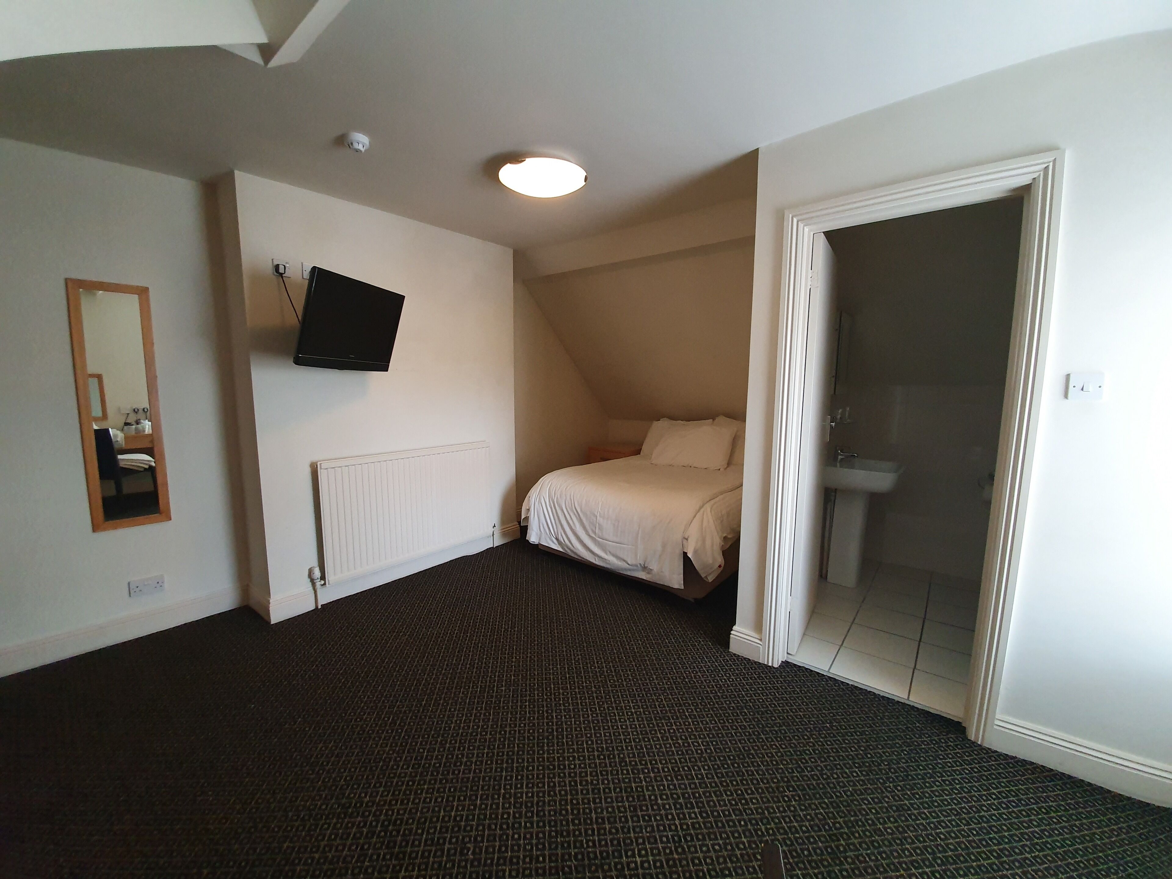 A bedroom with a bed, a mounted TV, a mirror, a radiator, and a door leading to a bathroom.