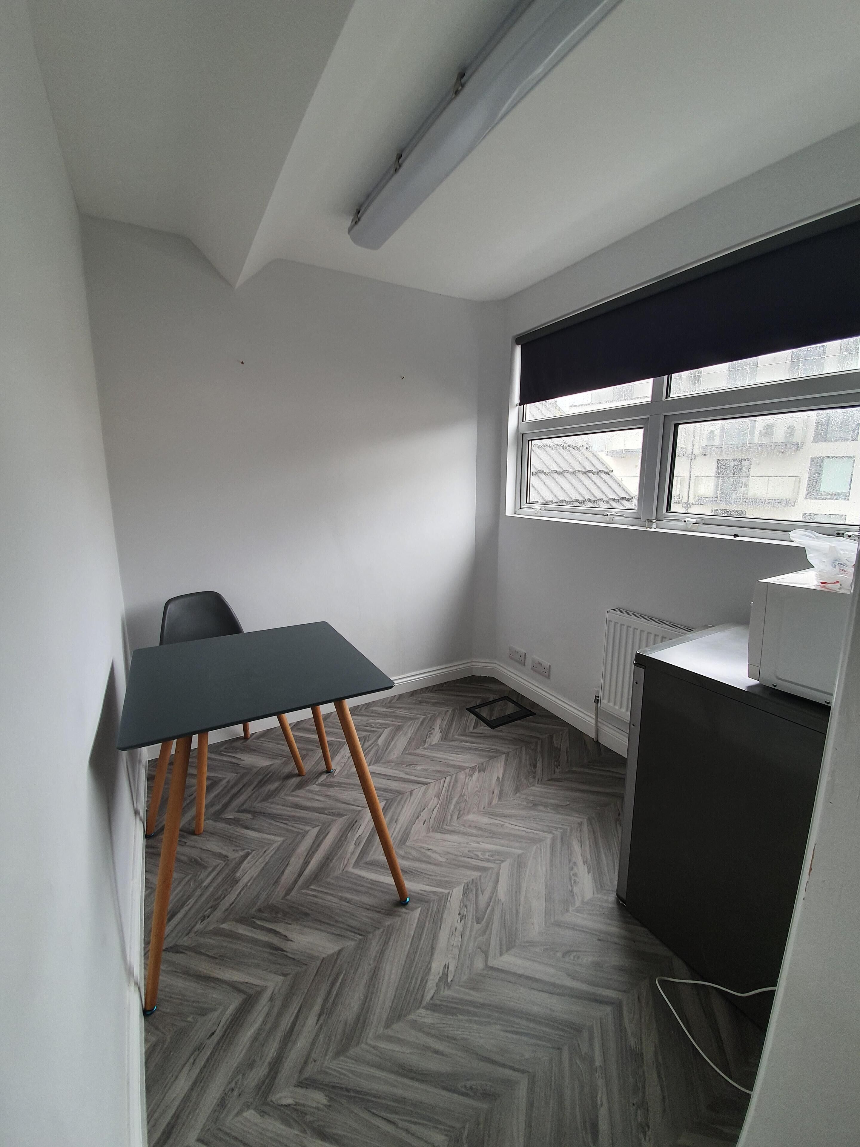 A small, modern room with a herringbone-patterned floor, a single table with wooden legs, a black chair, a radiator, and a window with a view of buildings outside.