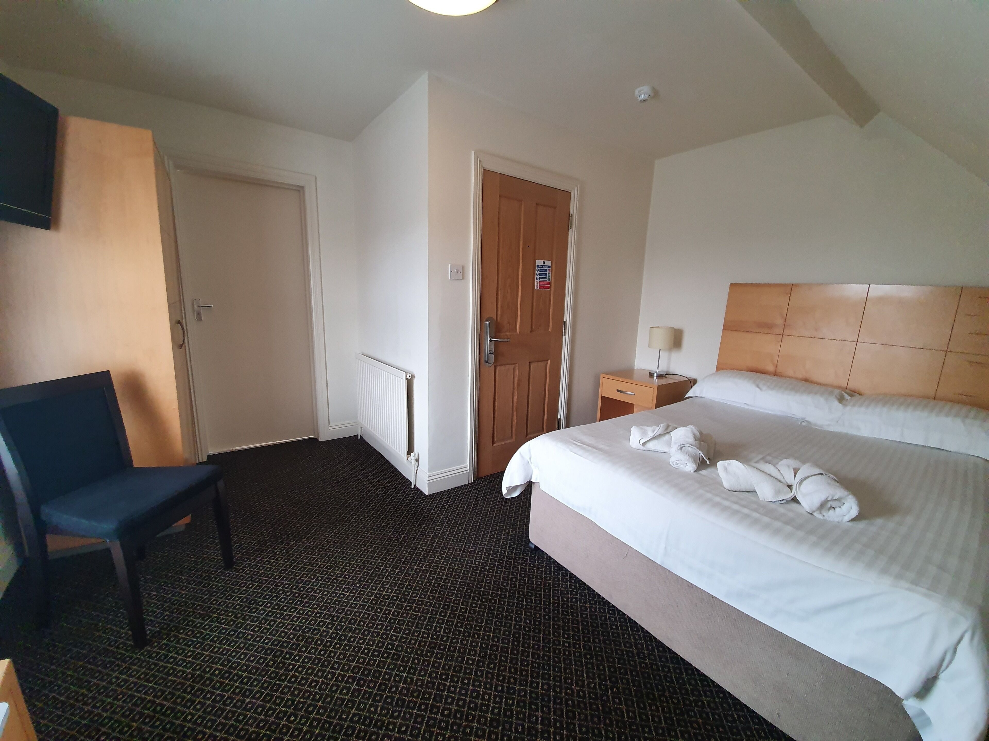 A hotel room with a bed, a chair, a bedside table with a lamp, and a radiator.