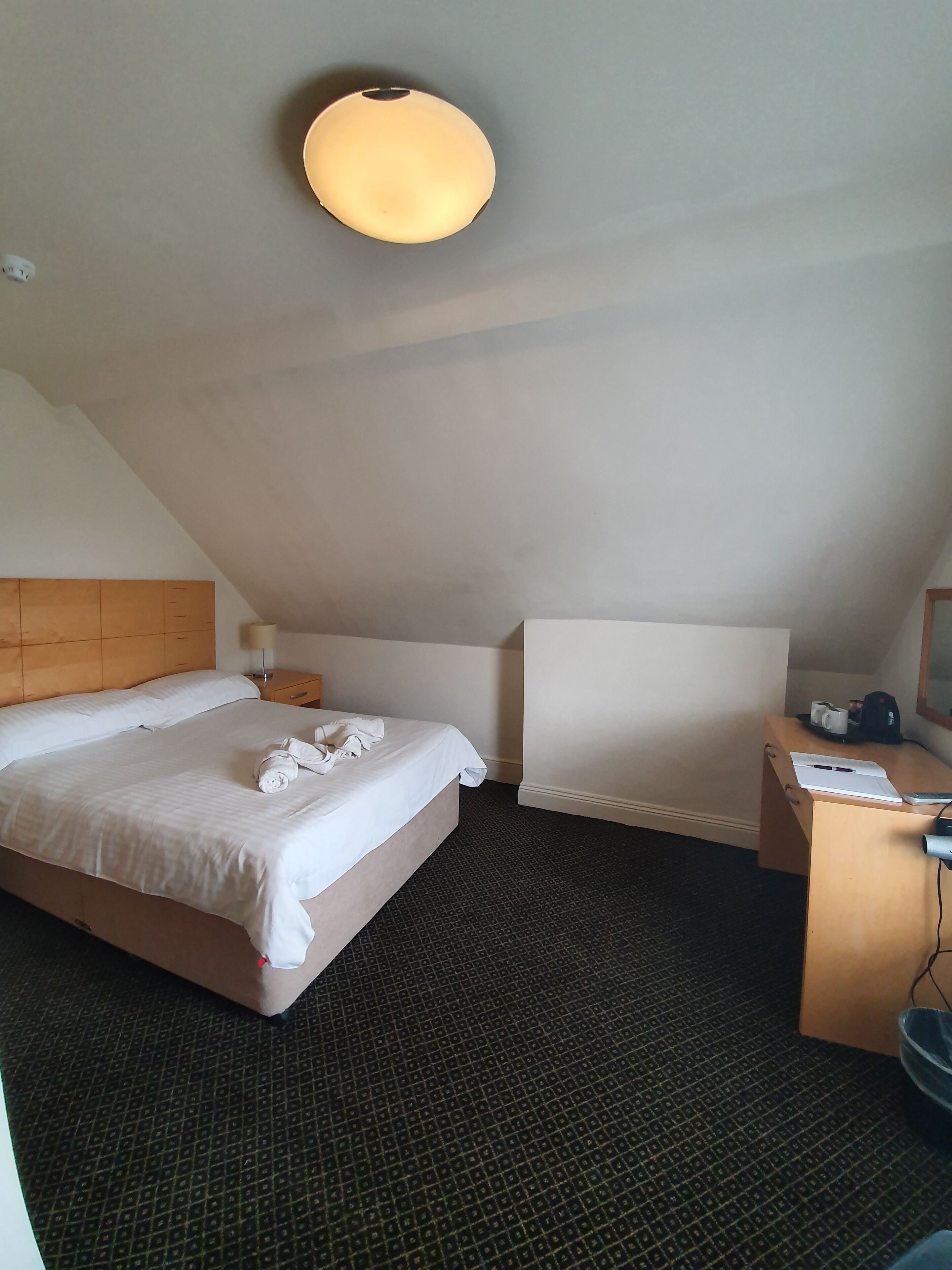 Quarto de hotel com cama, escrivaninha, cadeira, luminária e pequena mesa.