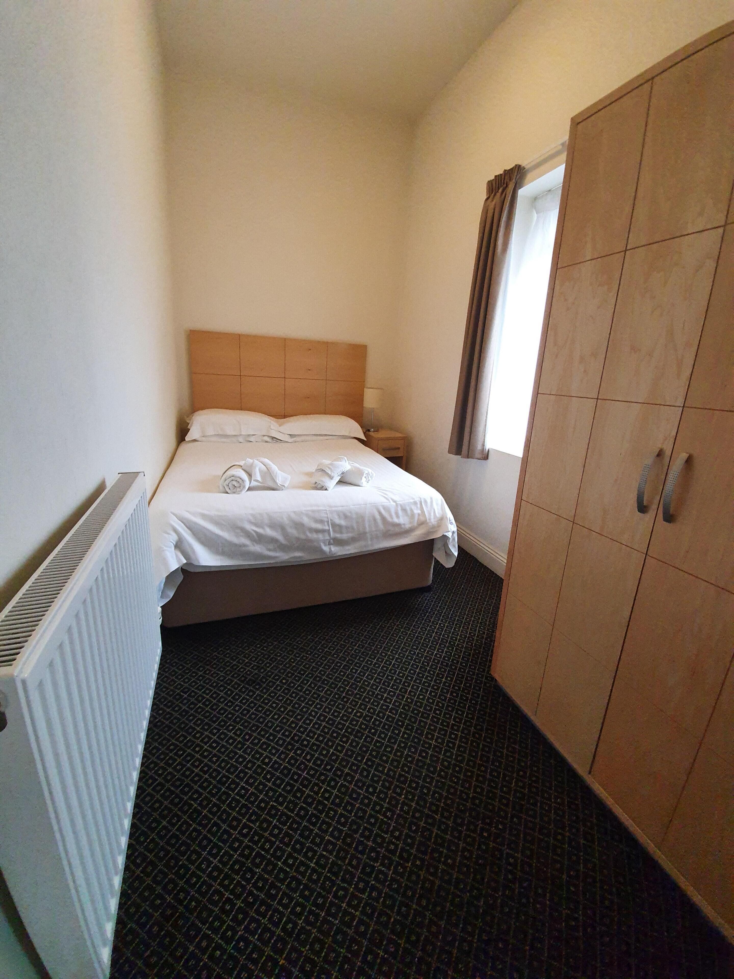 Une petite chambre d’hôtel propre, avec un lit simple, une armoire, un radiateur et une fenêtre avec des rideaux.
