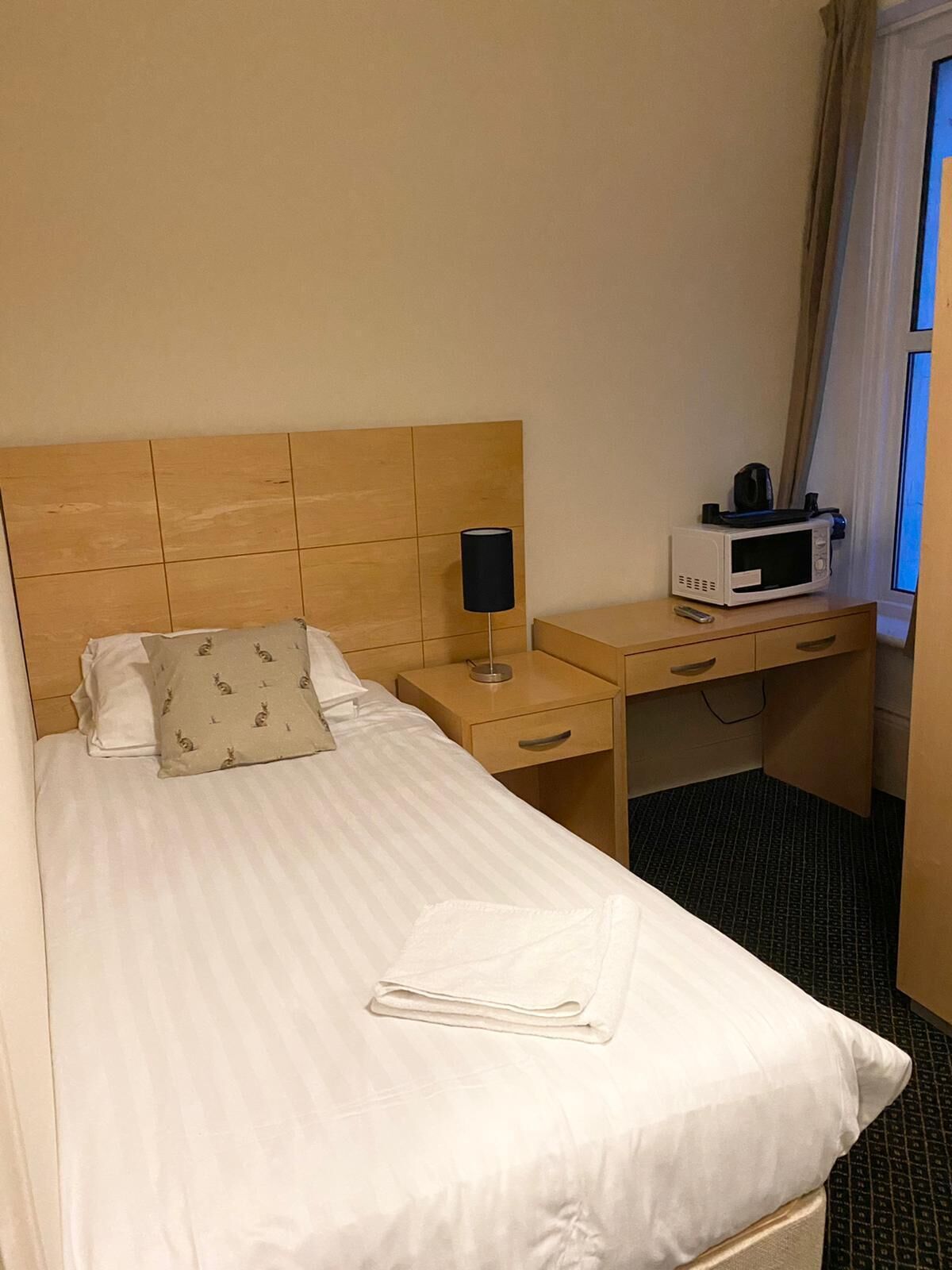 Habitación de hotel con cama, mesita de noche con lámpara, microondas y ventana con cortinas.