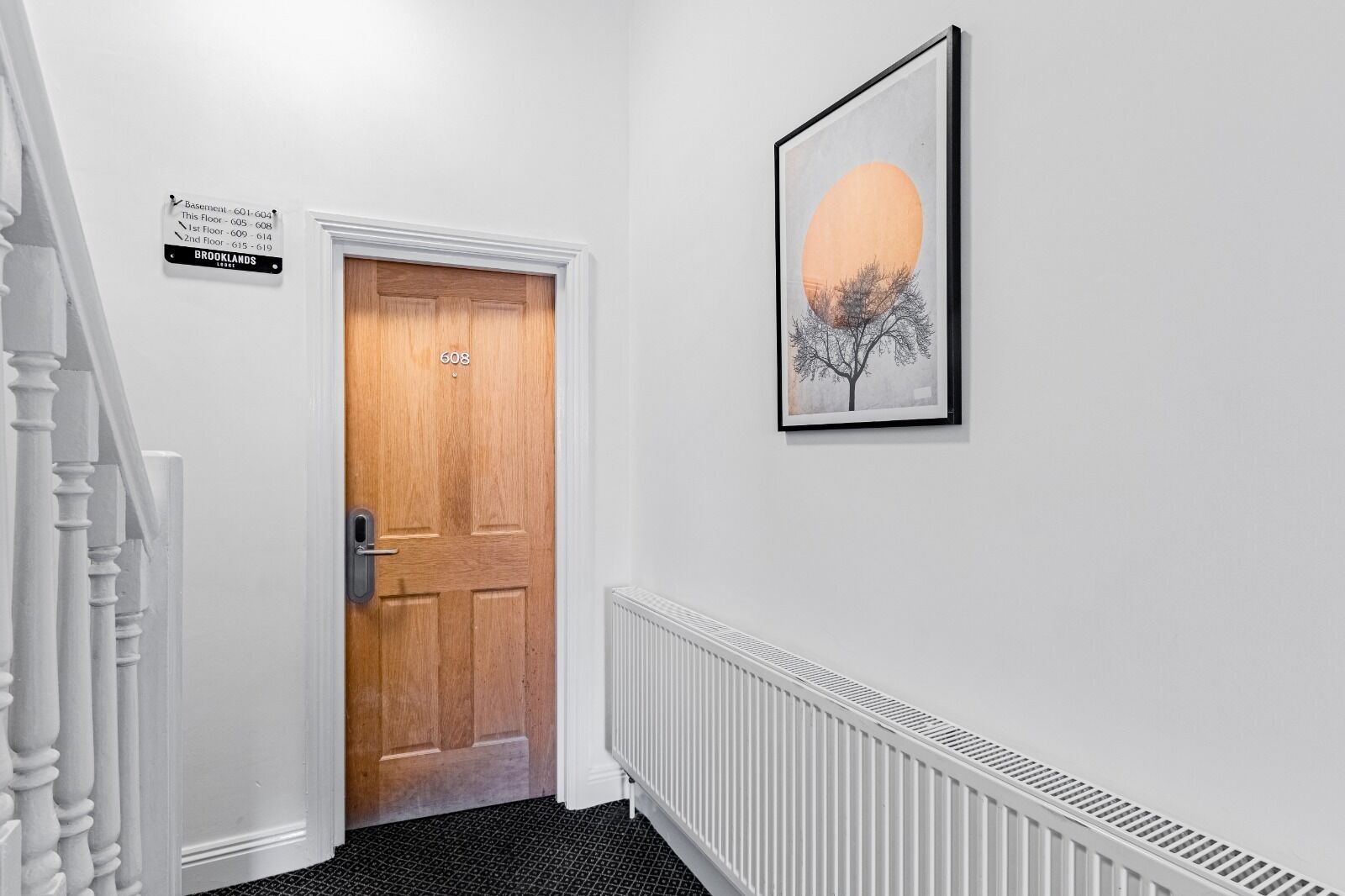 A hallway with a wooden door labeled 608, a sign indicating the basement and floor numbers, a radiator, and a framed picture of a tree against a sunset.