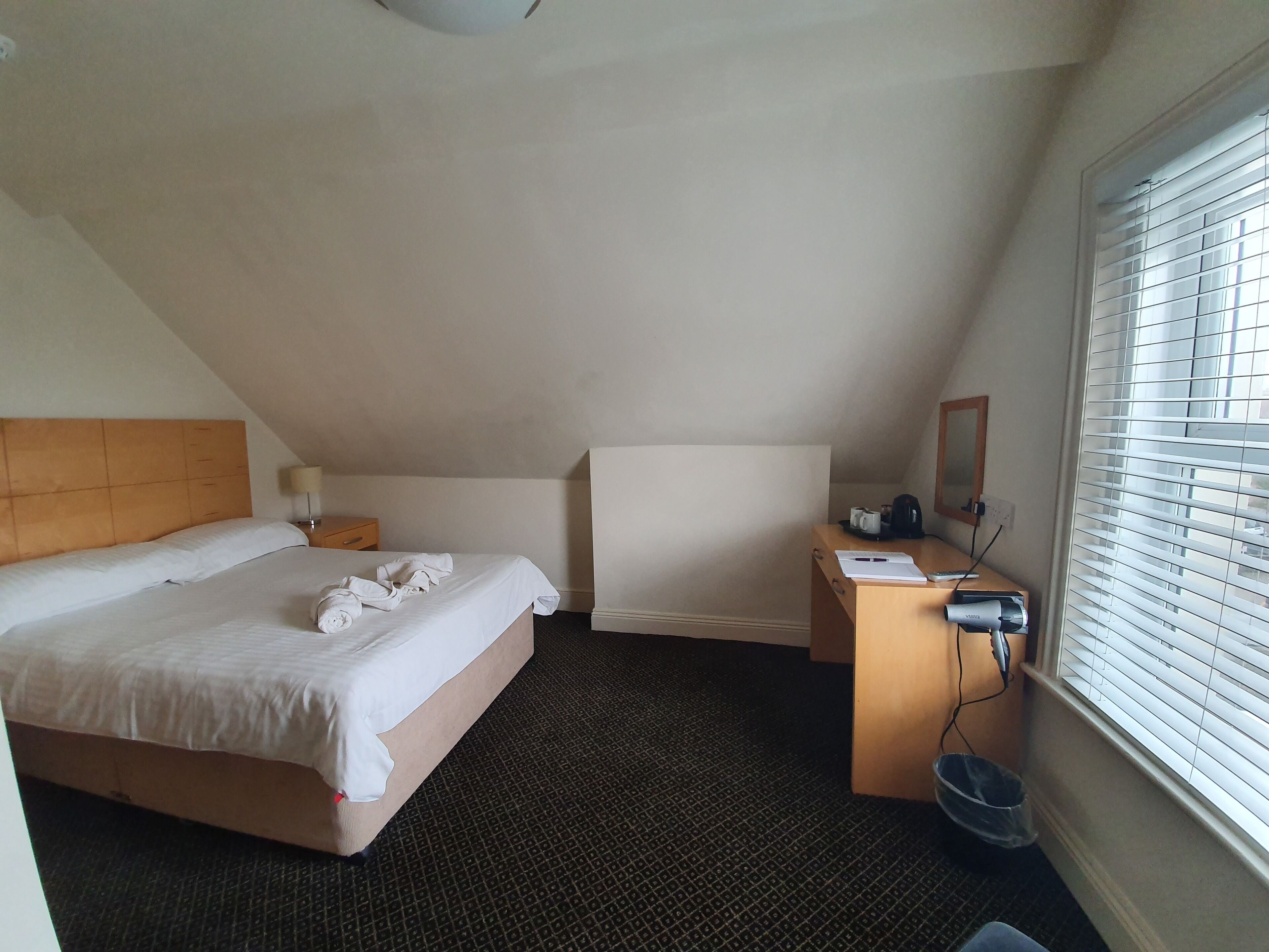 A hotel room with a bed, a desk, a chair, a window with blinds, and a small trash bin.