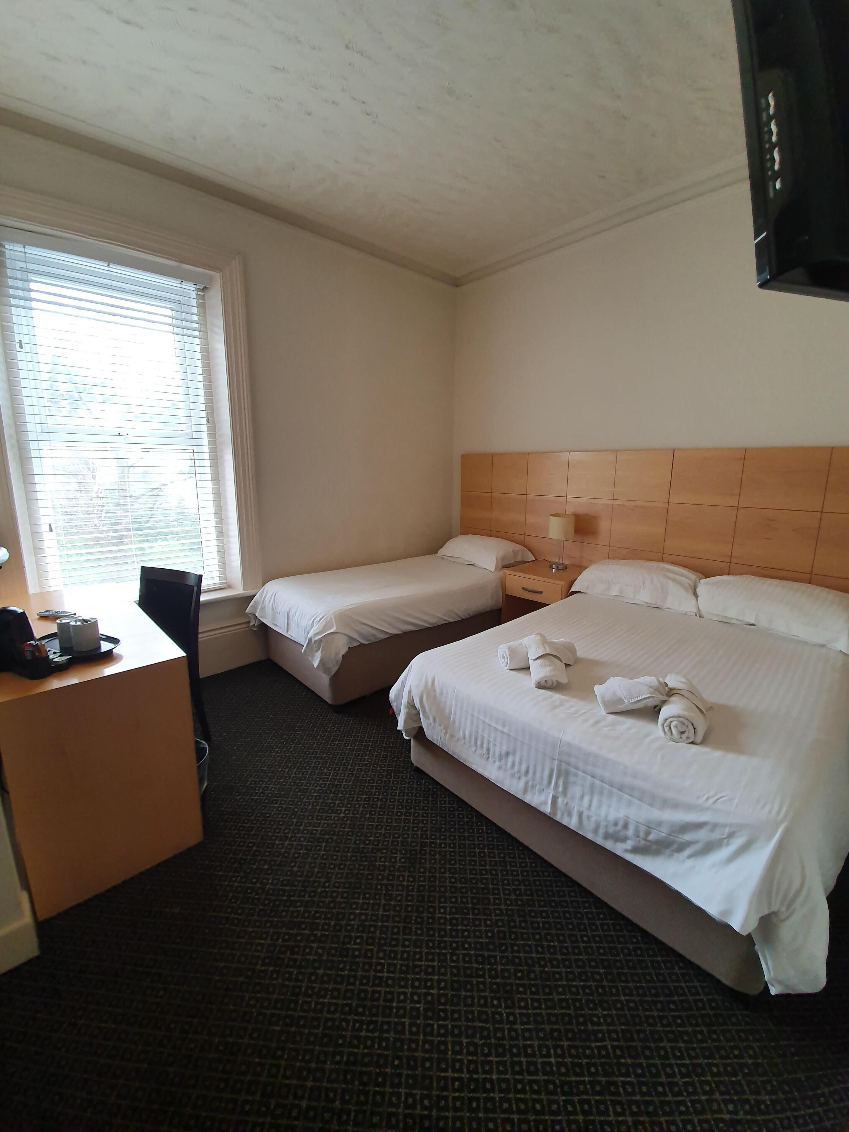 A hotel room with two single beds, a desk with a chair, a small table, and a window with blinds.