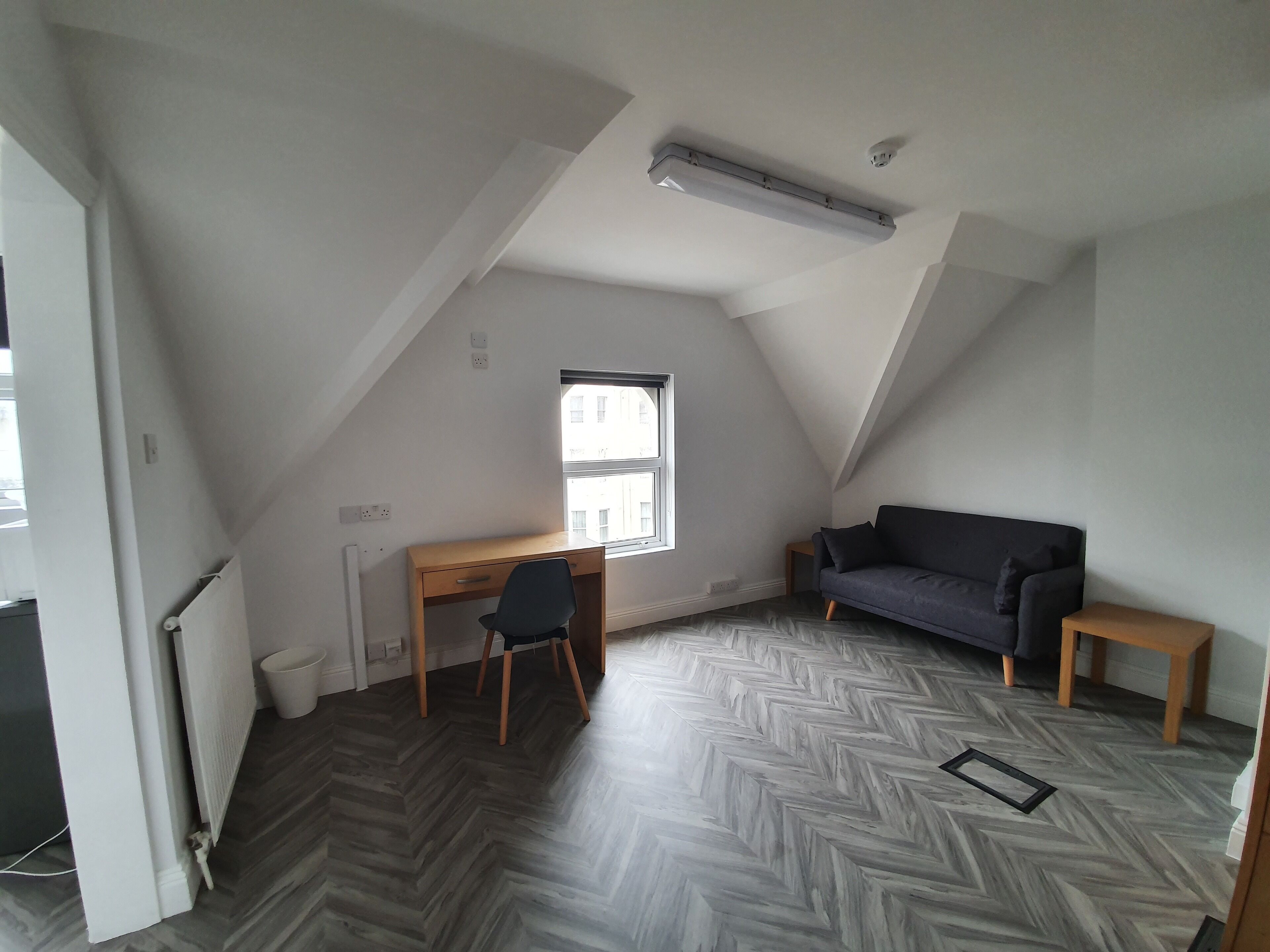 A room with a sloped ceiling, a desk with a chair, a dark sofa, and a small wooden table.