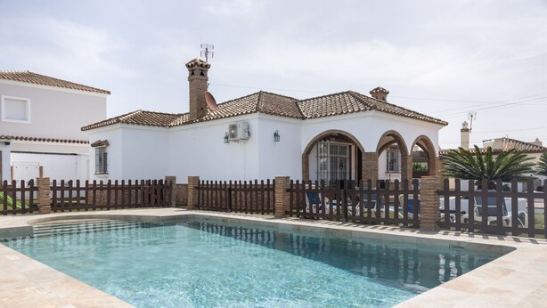 A swimming pool with a tiled deck, a white building with a tiled roof, and a wooden fence. - Family Home "La Lobita 1" Short Drive from Beach with Pool, Terrace and Wi-Fi (Conil de la Frontera)