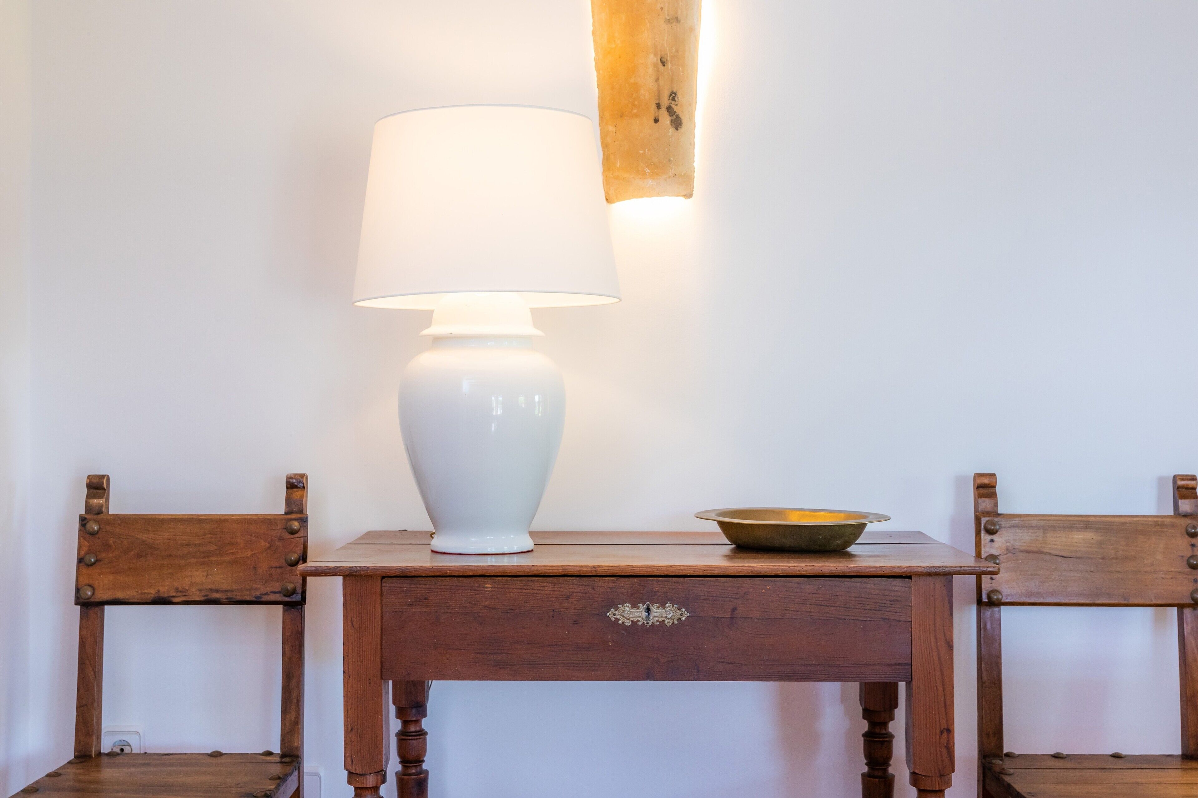 A wooden table with a white lamp and a bowl on it, flanked by two wooden chairs.