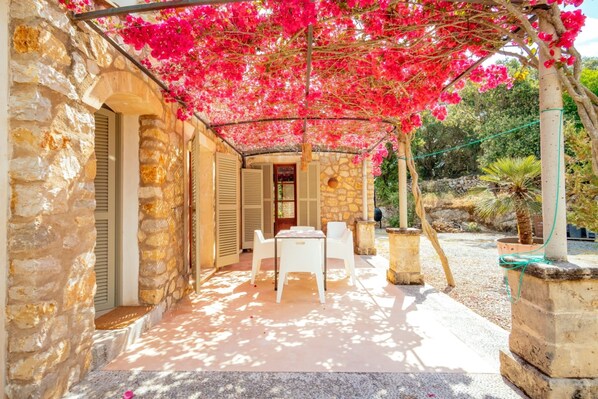 A stone building with a covered patio, white outdoor furniture, and a view of the surrounding garden.