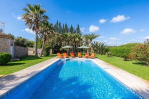 A swimming pool with lounge chairs and umbrellas, surrounded by palm trees and a stone building.