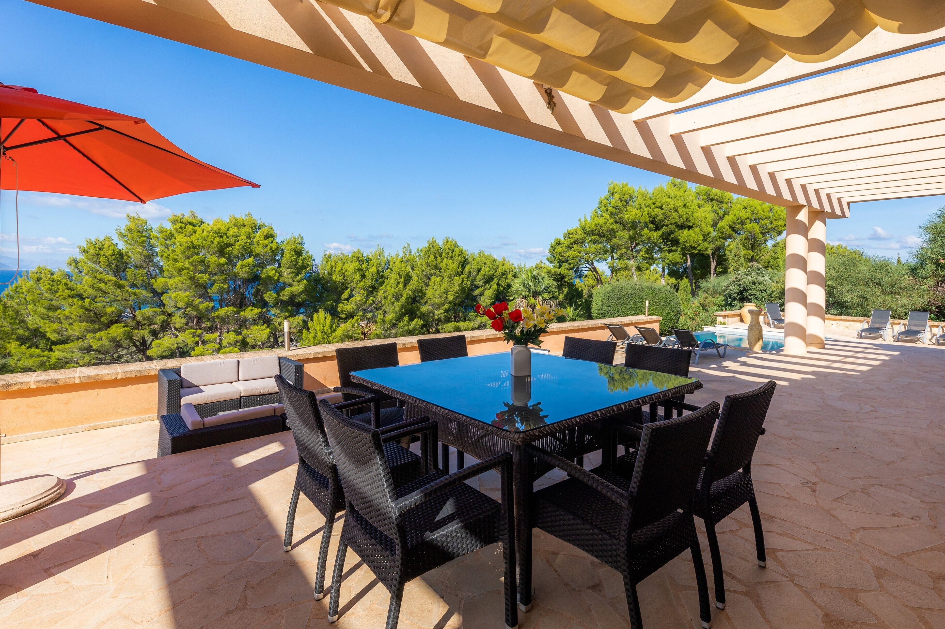 Een terras buiten met een tafel, stoelen, een rode parasol en uitzicht op bomen en de zee.