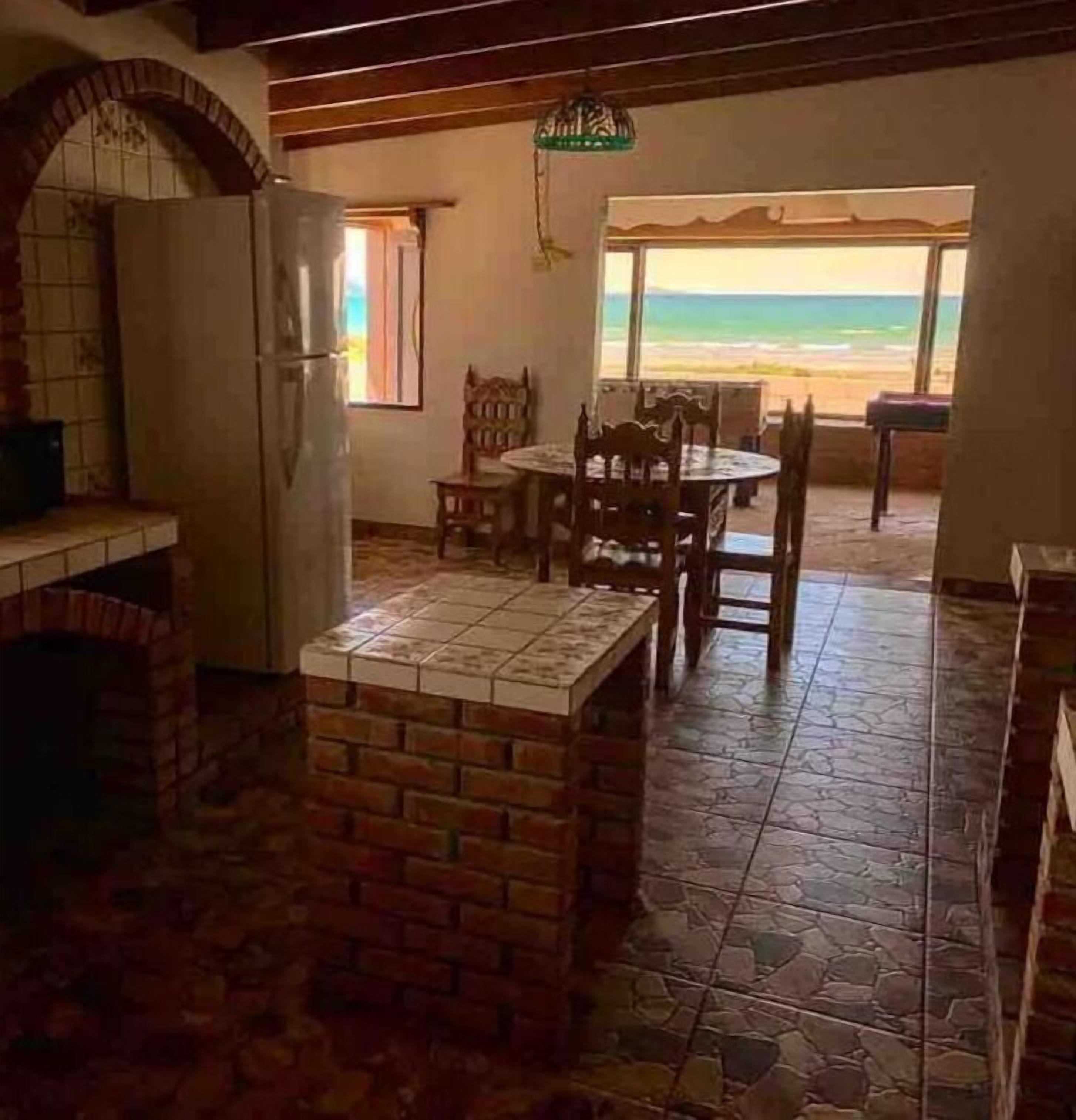 A kitchen with a brick island, a dining table with chairs, a refrigerator, and a view of the beach.