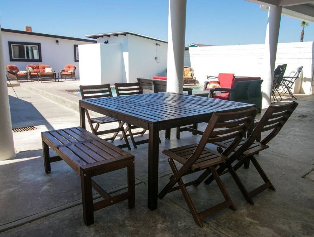 An outdoor patio area with a wooden table and chairs, a small white building, and a white wall in the background.