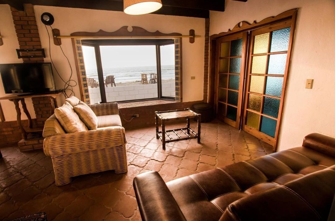 A living room with a sofa, a wicker chair, a small table, a TV, and a large window with a view of the beach.