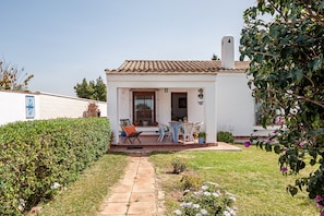 A white building with a tiled roof, a patio with chairs and a table, and a pathway leading to the entrance.