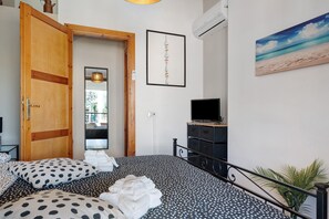 A bedroom with a bed, a wooden door, a framed artwork, an air conditioning unit, and a television.