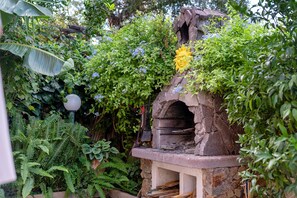 A stone-built outdoor oven with a chimney, surrounded by lush greenery and flowering plants. - Charming Villetta Lelù with Garden and Terrace by the Sea (Villasimius)