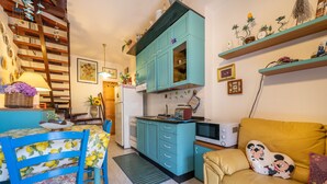 A compact kitchen with teal cabinets, a dining table with chairs, a sofa with a Mickey Mouse pillow, and a staircase. - Charming Villetta Lelù with Garden and Terrace by the Sea (Villasimius)