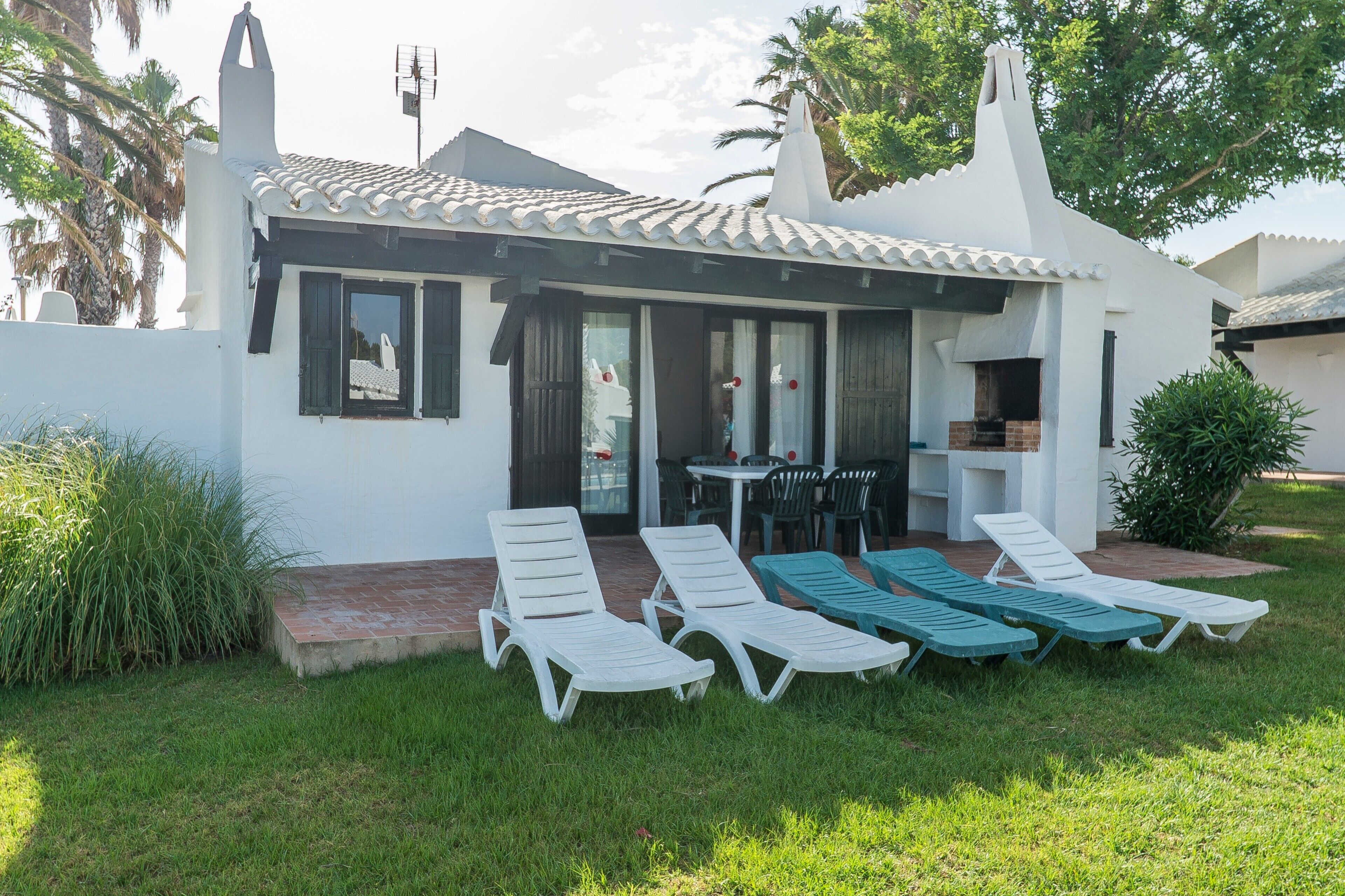 A small house with a tiled roof, a patio with lounge chairs, and a table.