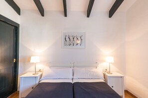 A double bed with white linens, two bedside tables with lamps, a framed artwork above the bed, and dark wooden beams on the ceiling.