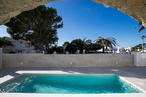 Ett poolområde med en klarblå pool, omgivet av en låg stenmur, med träd och byggnader i bakgrunden.