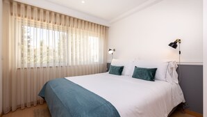 A neatly made bed with a blue and white bedspread, a window with sheer curtains, and two wall-mounted lamps. - Feel Corporate Housing Marquês II (Oporto)