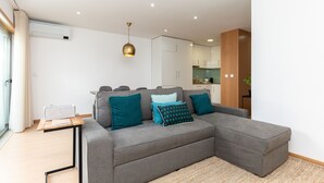 A modern living room with a grey sectional sofa, teal cushions, a small wooden side table, and a hanging pendant light. - Feel Corporate Housing Marquês II (Oporto)