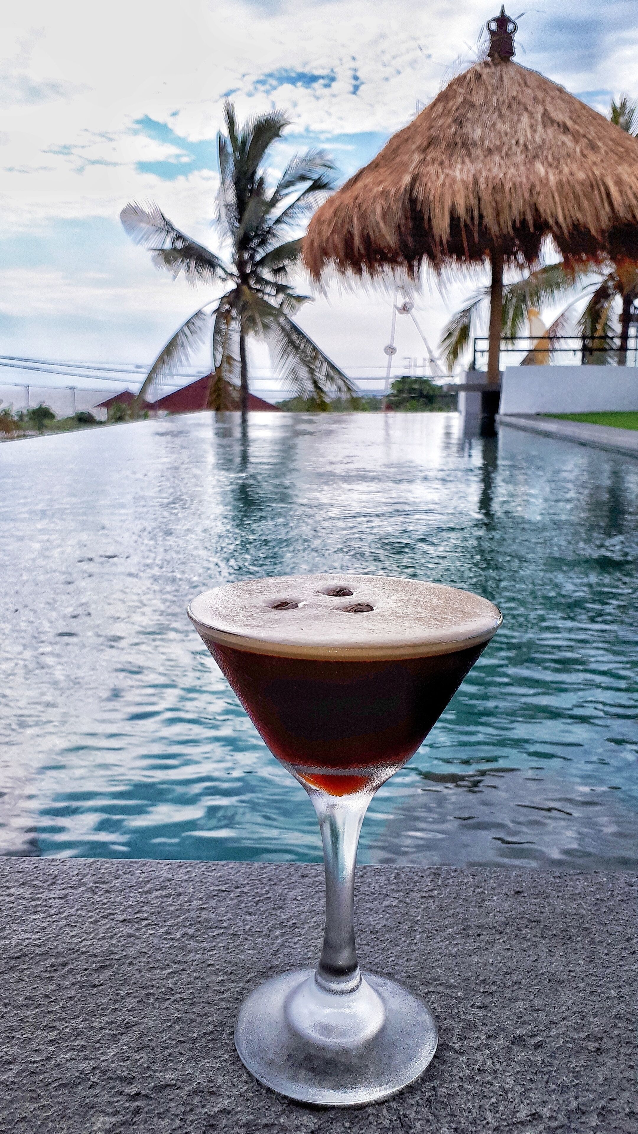 Een cocktail in een martini glas, met een zwembad en palmbomen op de achtergrond.