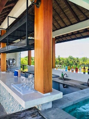 Zona de bar al aire libre con piscina, pilares de madera y techo de paja.