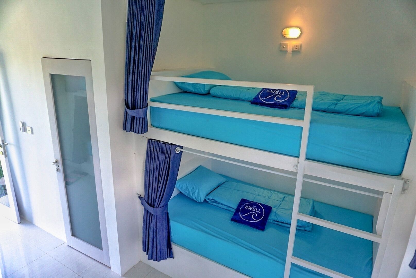 Una cama litera con ropa de cama y almohadas azules, una puerta con un panel de vidrio y un aplique fijado a la pared.