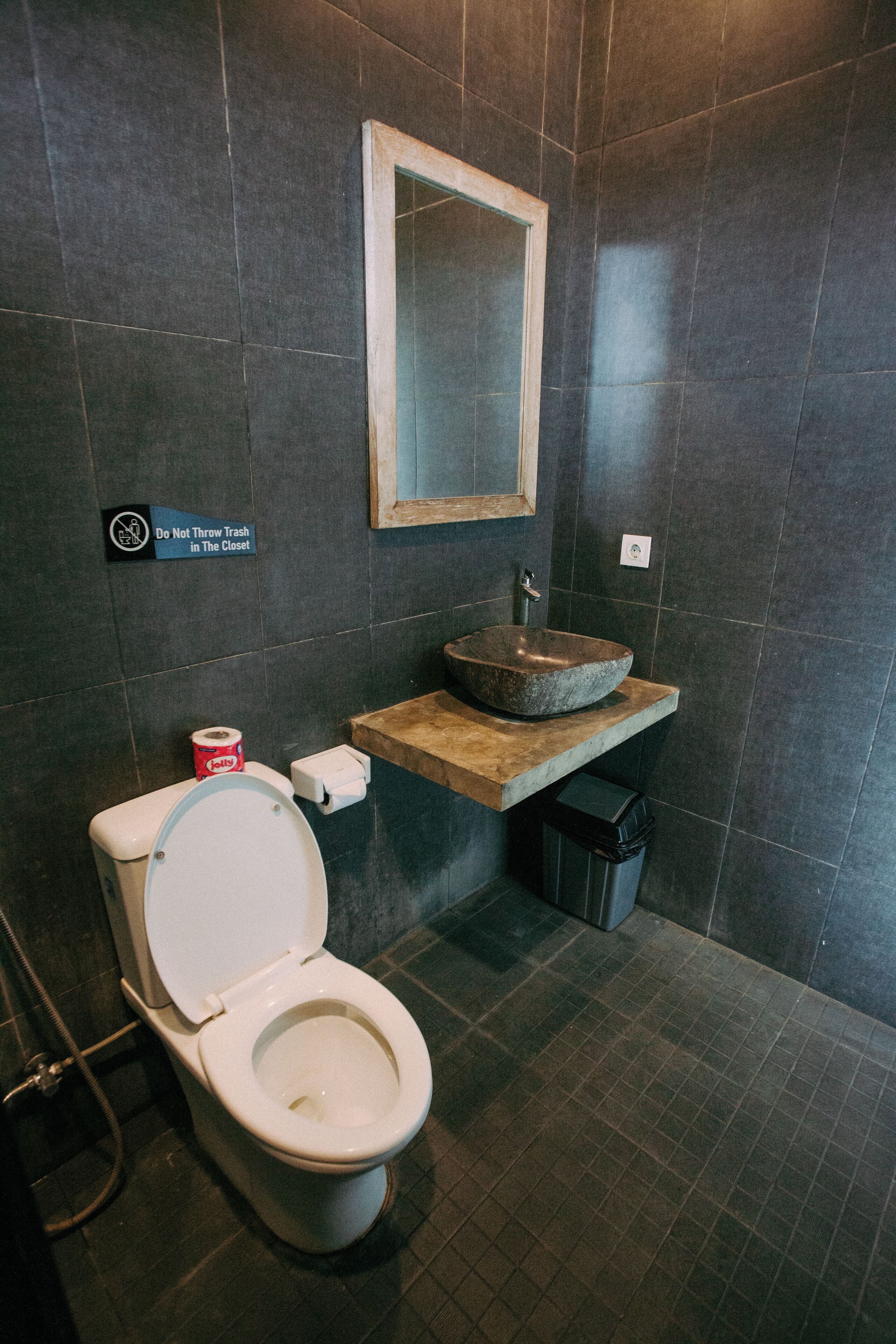 Ein Badezimmer mit Toilette, Waschbecken und Spiegel. Auf der Wand ist ein Schild mit der Aufschrift 'Keinen Müll in den Schrank werfen.'
