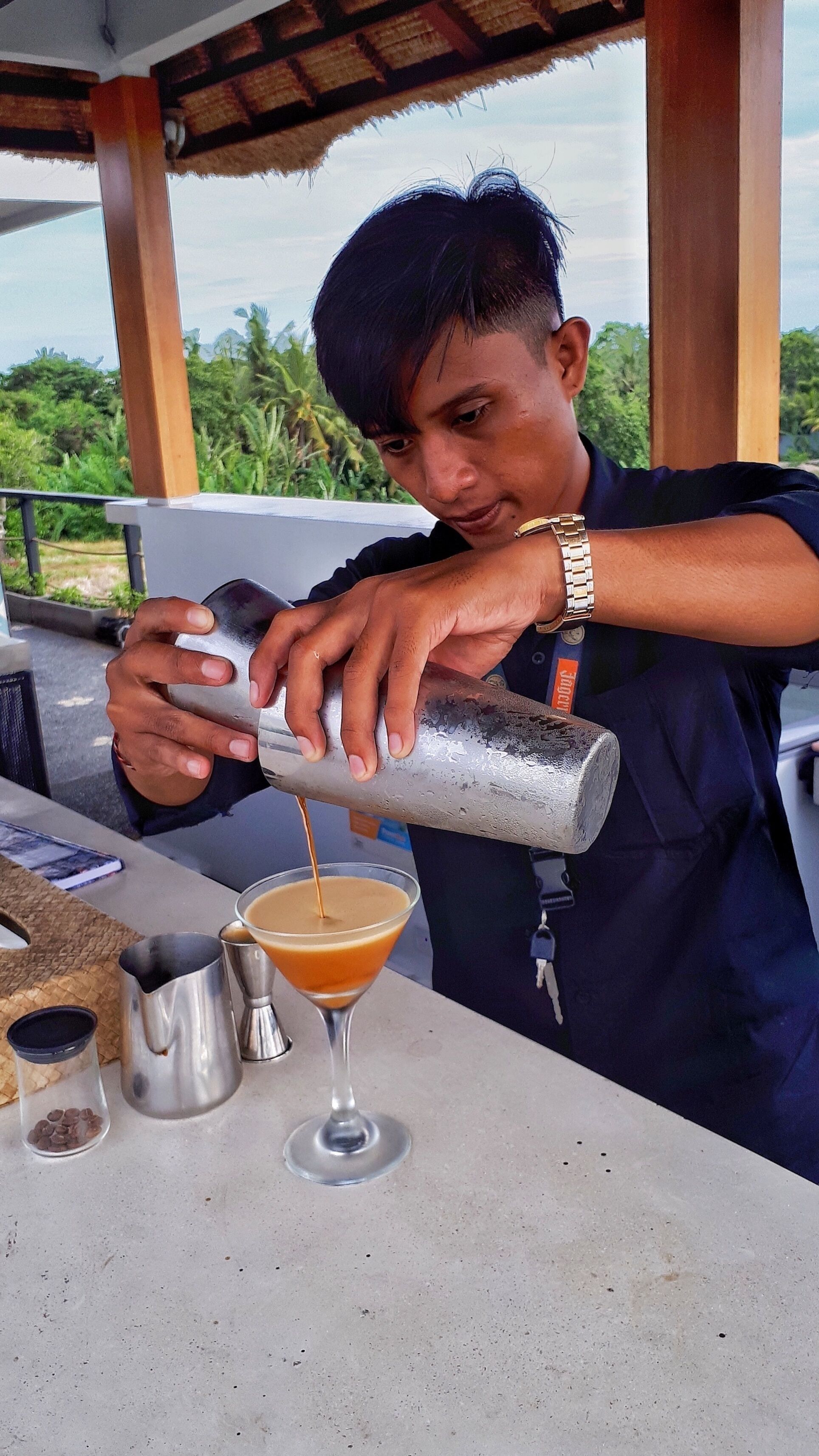 A person pouring a drink into a martini glass at a bar with a tropical view.