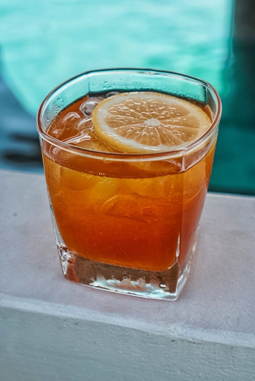 Een glas amberkleurig drankje met een stukje citrusvrucht op de rand, geplaatst op een oppervlak met een blauwgroene achtergrond.