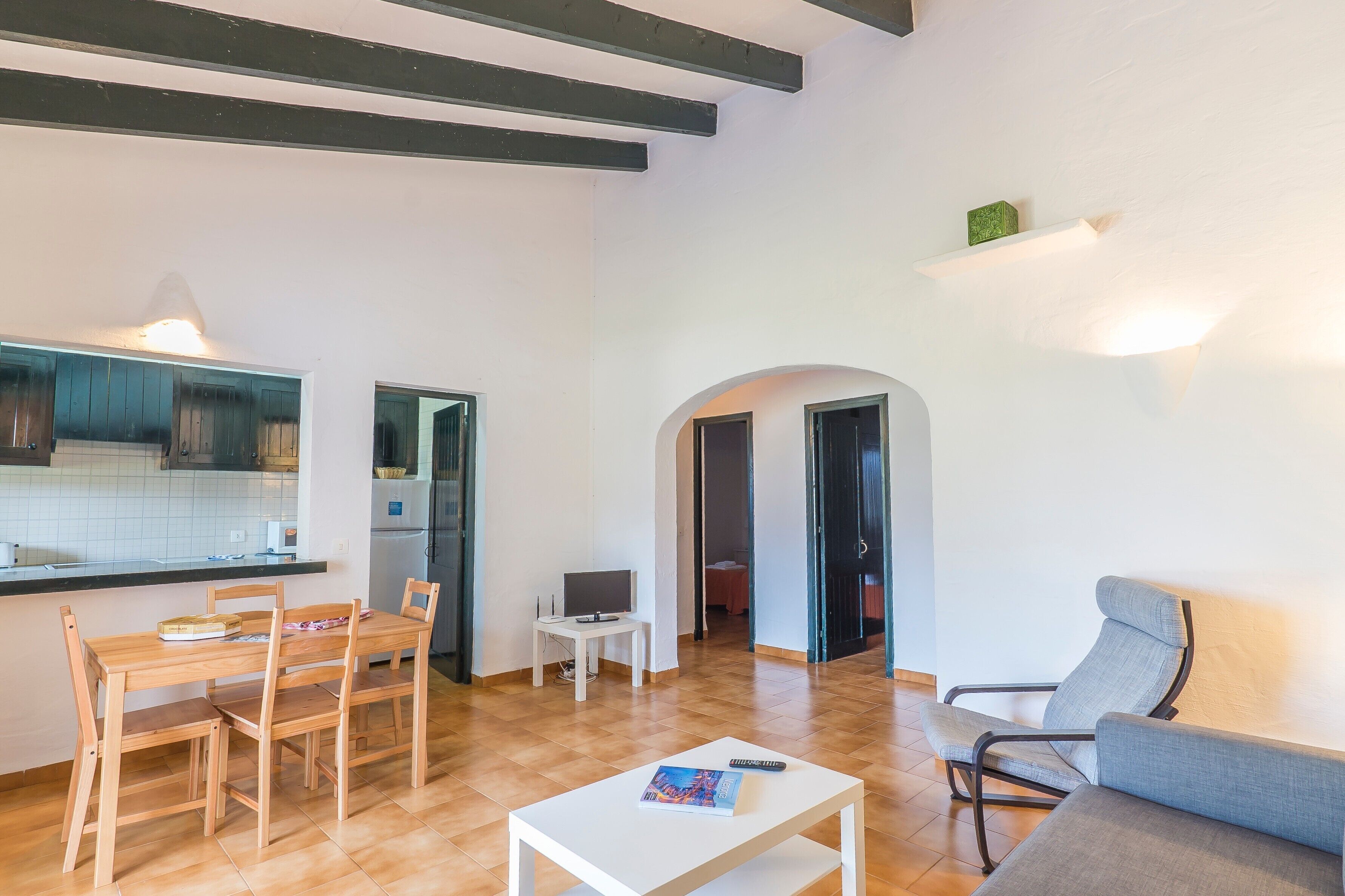 Una sala de estar con un sofá, una mesa de comedor con sillas, una pequeña mesa blanca y un televisor. Se observa un área de cocina con una encimera y alacenas.