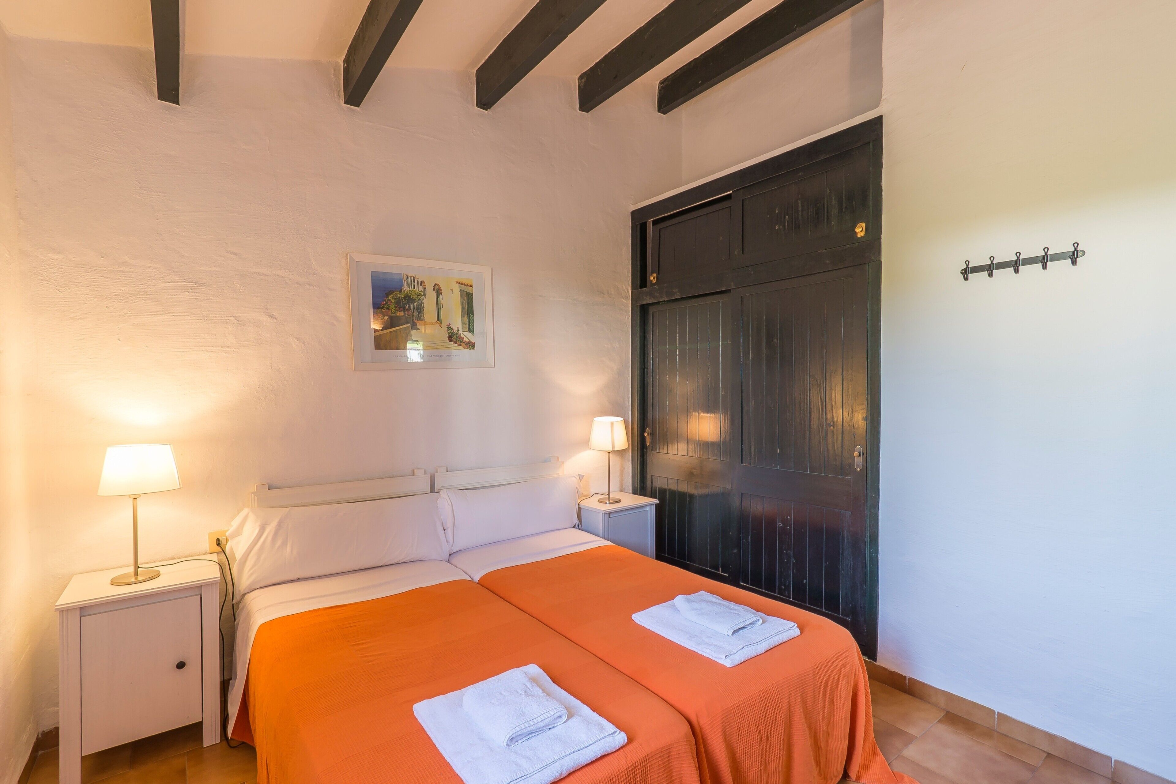 A double bed with an orange bedspread, a nightstand with a lamp, a painting on the wall, and a dark door.