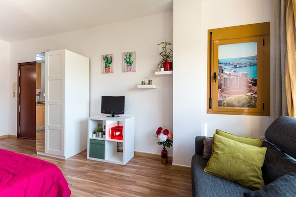 A room with a bed, a TV on a white stand, a sofa with a green pillow, a wooden door, and a cityscape painting on the wall.