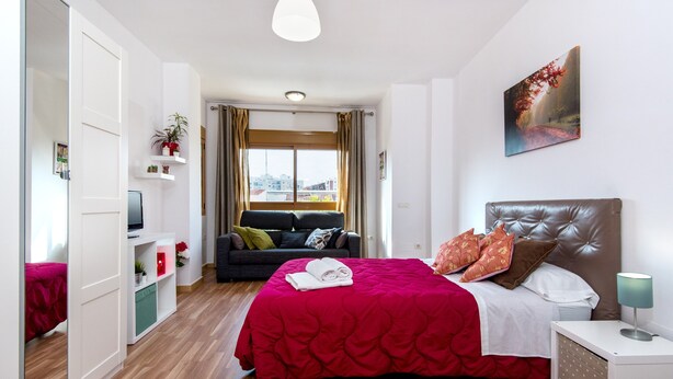 Habitación de hotel con cama, mesita de noche, televisión, ventana con cortinas, sofá y vistas a la ciudad.