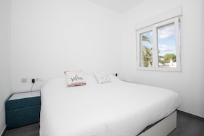 Un dormitorio con una cama, una mesita de noche, una ventana que ofrece vistas a palmeras y un enchufe de pared.
