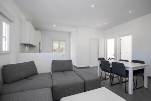 A modern living room with a grey sofa, a dining table with chairs, and a kitchen area with white cabinets and a window.
