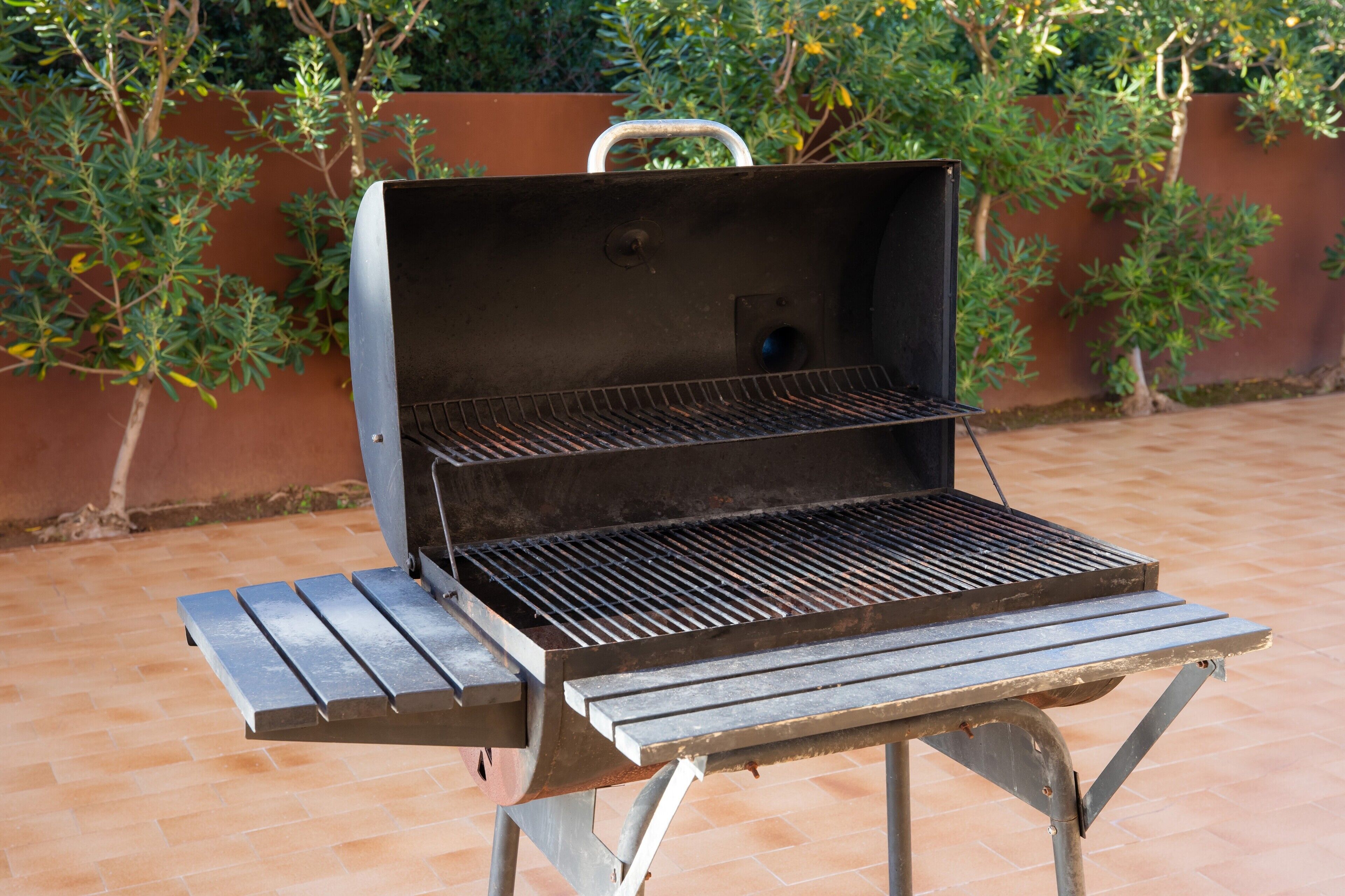 Ein tragbarer Grill mit Metallrost und Tragegriff, auf einer gefliesten Terrasse mit Backsteinmauer und Grünpflanzen im Hintergrund.