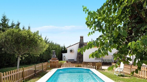 Une piscine rectangulaire entourée d’une clôture en bois, avec un bâtiment blanc et une cheminée en arrière-plan.