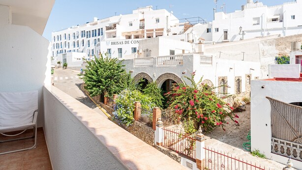 A balcony with a white chair, a stone wall, and a view of white buildings with arches and balconies.