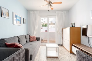 Een moderne woonkamer met een grijze bank, een kleine witte tafel, een houten kast en een plafondventilator.
